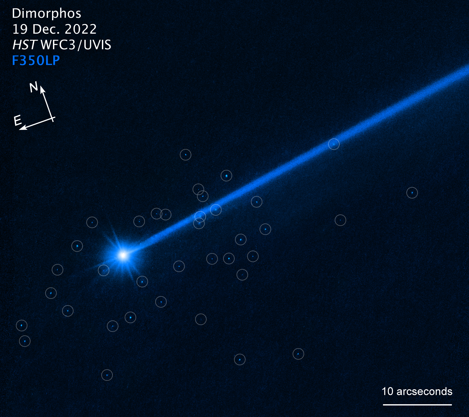 Asteroid biru terang dengan ekor panjang mengarah ke kanan atas.  Batu-batu biru kecil dilingkari di sekitar asteroid