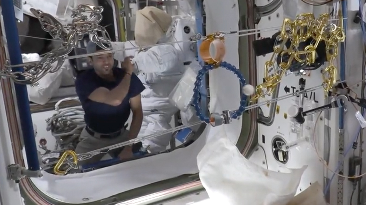 astronot mengambang di modul stasiun ruang angkasa.  dia menghadapi dua tali pengikat yang direntangkan di pintu masuk.  di antara kedua tali itu ada dua cincin kecil, satu di atas yang lain.  dia melempar bola melalui salah satu ring.