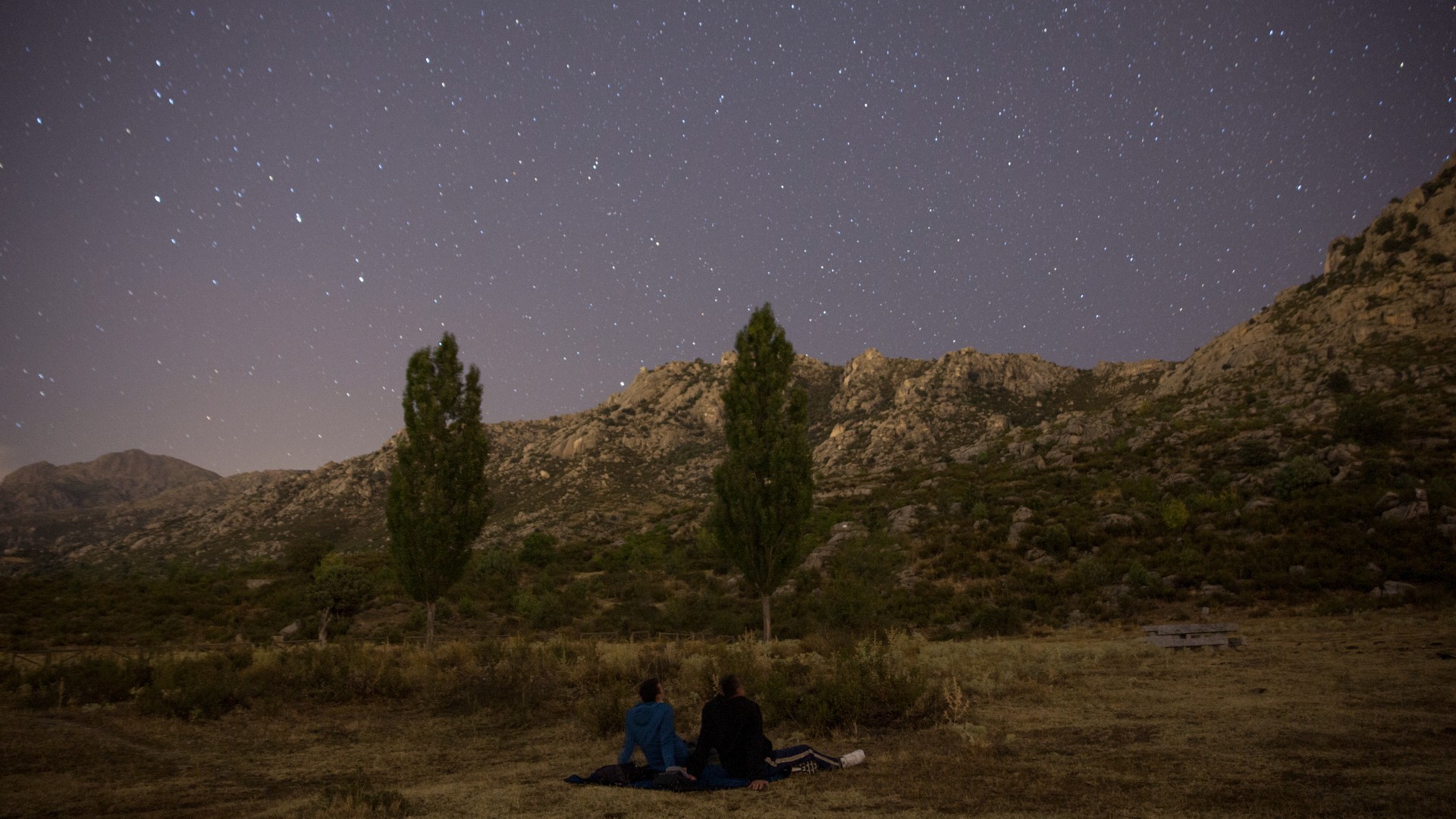 two people sit on a blanket in a field under the stars