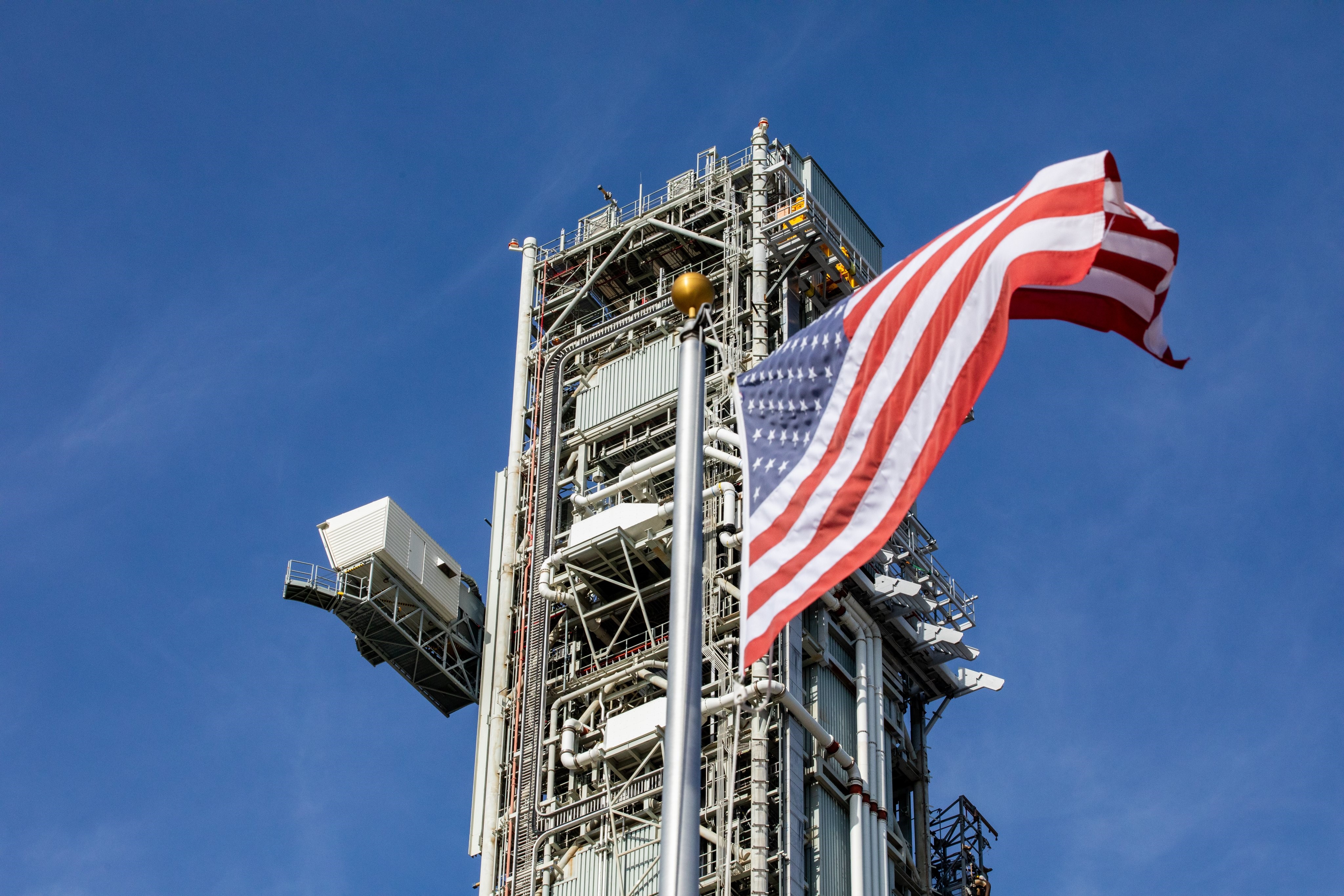 Peluncur seluler 1 terlihat di latar belakang sementara bendera Amerika berkibar tertiup angin di latar depan.  Perayap membawa peluncur seluler 1 dari lokasi taman ke Launch Pad 39B.  Langit sebagian berawan dan biru.  Peluncur seluler 1 memiliki lengan akses kru yang terlihat di sebelah kiri dan banyak potongan rumit terlihat di seluruh bagiannya.