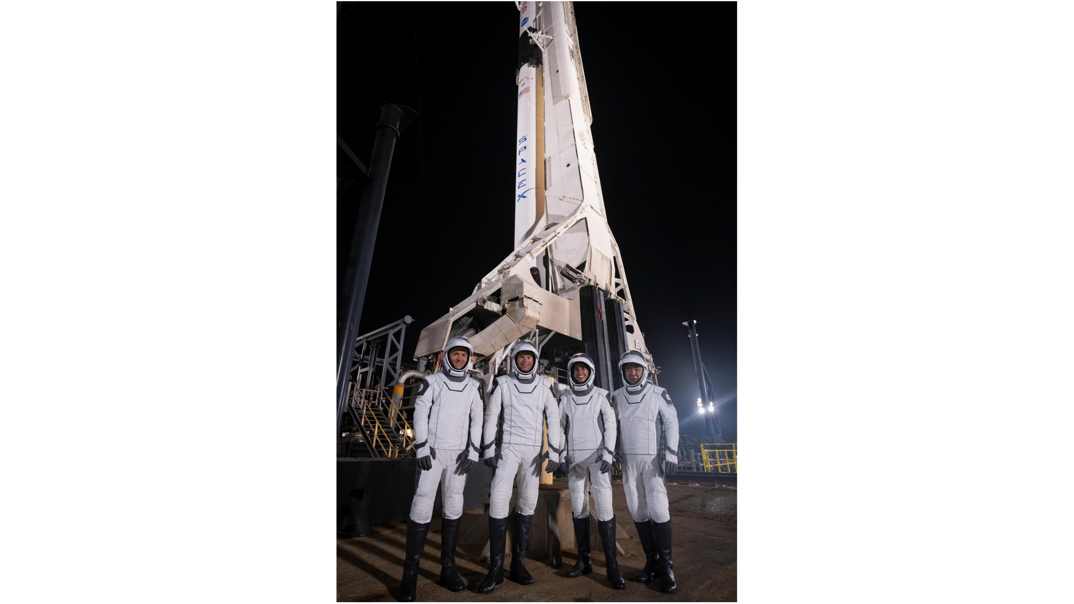 empat astronot dan pakaian antariksa putih dan hitam berdiri di depan roket di landasan peluncuran pada malam hari.