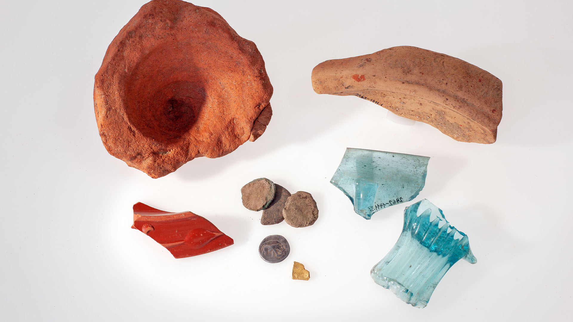 Alas amphora, potongan lesung, tepi mangkuk, empat koin, sebuah benda emas, potongan botol, dan mangkuk kaca biru.