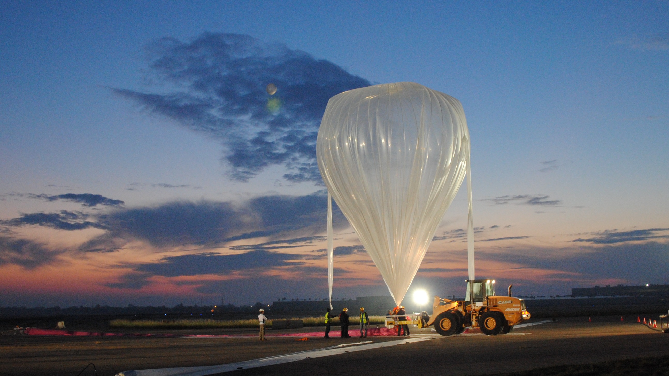 balon stratosfer yang mengembang sambil diikat ke tanah.  sebuah kendaraan pendukung kecil berdiri di sebelah kanan balon.  di belakang adalah langit saat fajar, dengan langit oranye dan biru serta sedikit awan