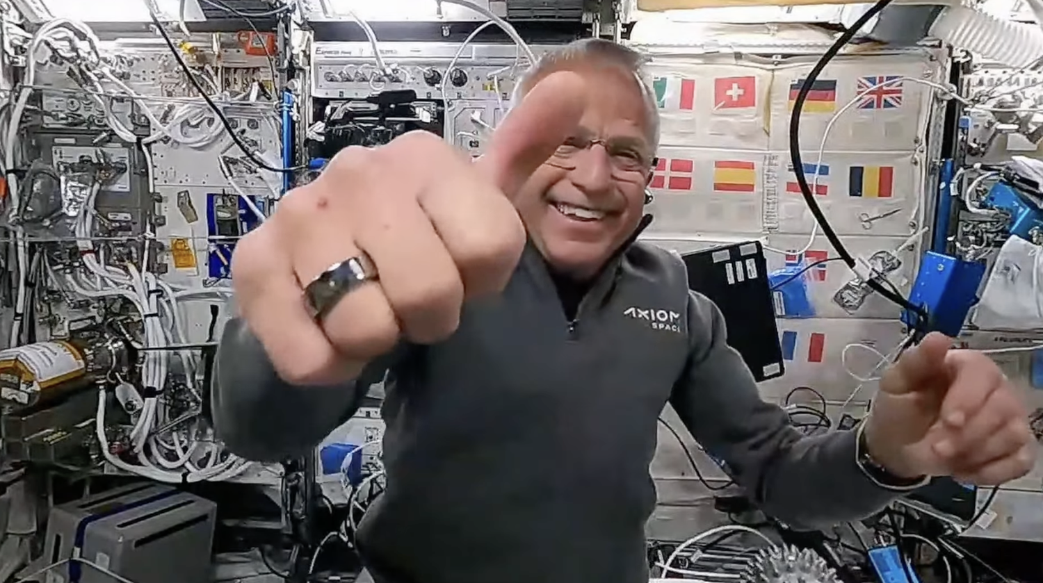 seorang astronot mengacungkan jempol ke kamera di luar angkasa.  tangan kanan sangat dekat dengan kamera dan terlihat cincin di jari telunjuk.  Di belakang, sang astronot tersenyum lebar sambil mengenakan kemeja bertuliskan 