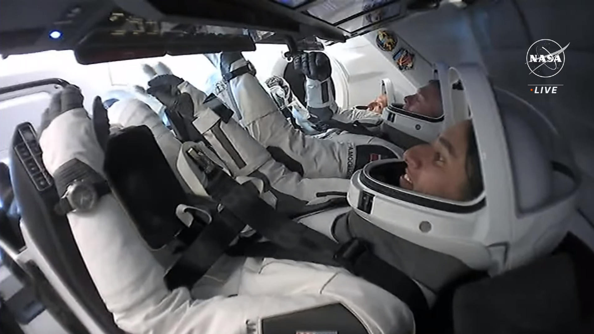 tiga astronot dalam pakaian antariksa putih dengan pelat muka helm terbuka duduk di dalam kapsul SpaceX di orbit