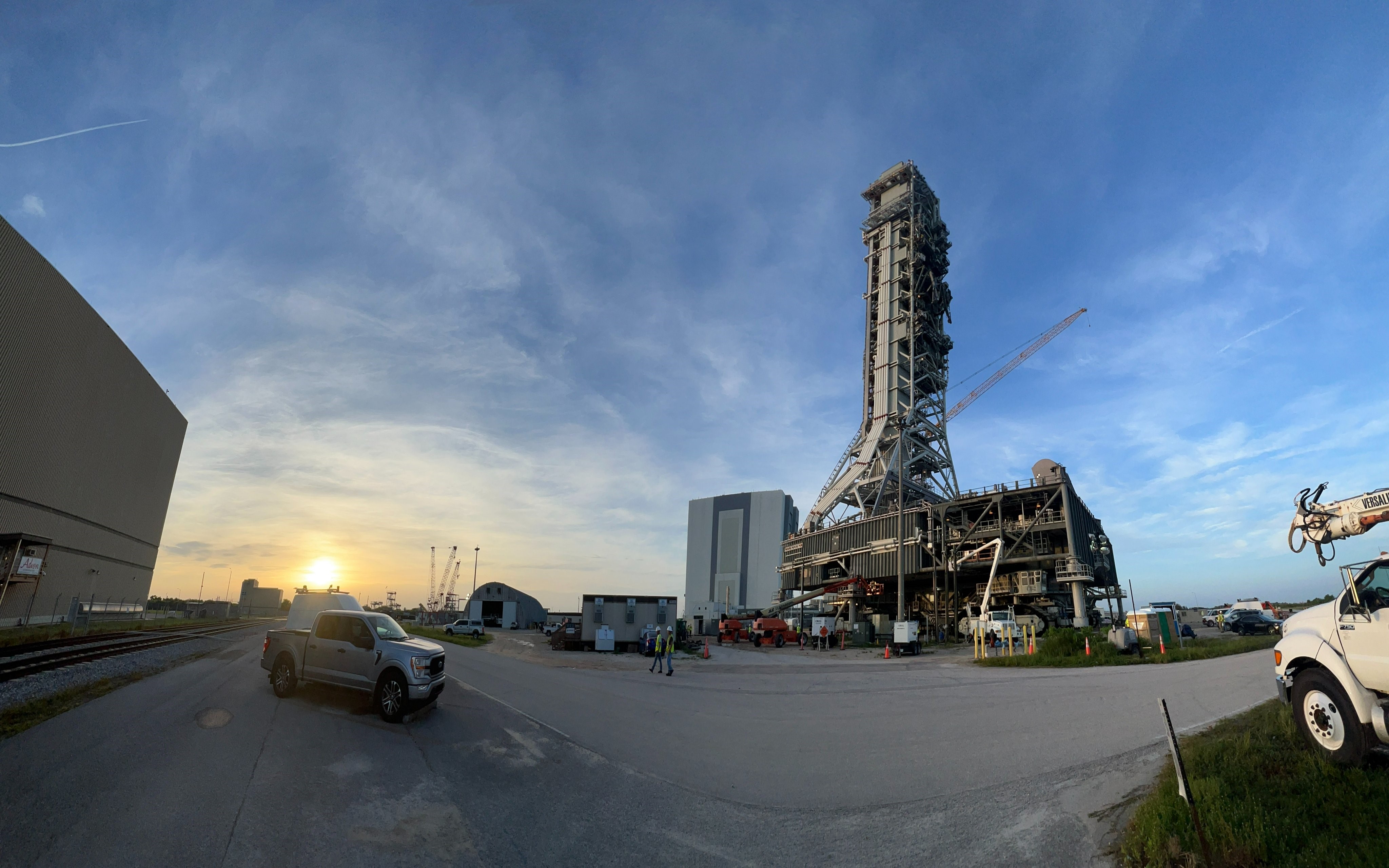 Matahari terbit, di sebelah kiri, menyinari langit biru di Kennedy Space Center NASA.  Di latar depan, kanan, adalah peluncur seluler 1 yang menjulang tinggi di atas tanah.  Gedung Perakitan Kendaraan terlihat di belakang.