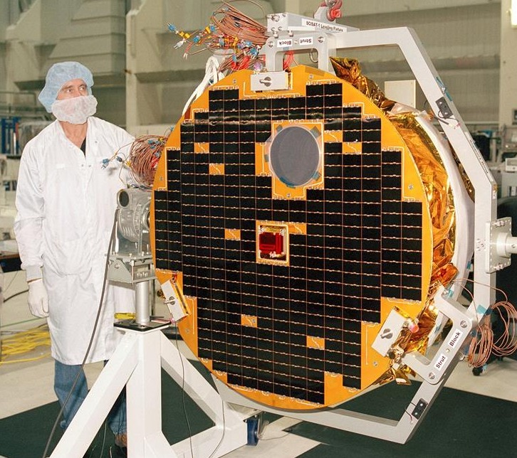 satelit melingkar dengan panel surya hitam di laboratorium.  tingginya hampir sama dengan teknisi yang berdiri di sebelah kirinya.  teknisi mengenakan jas lab, jaring rambut, dan masker untuk melindungi satelit dari kontaminasi