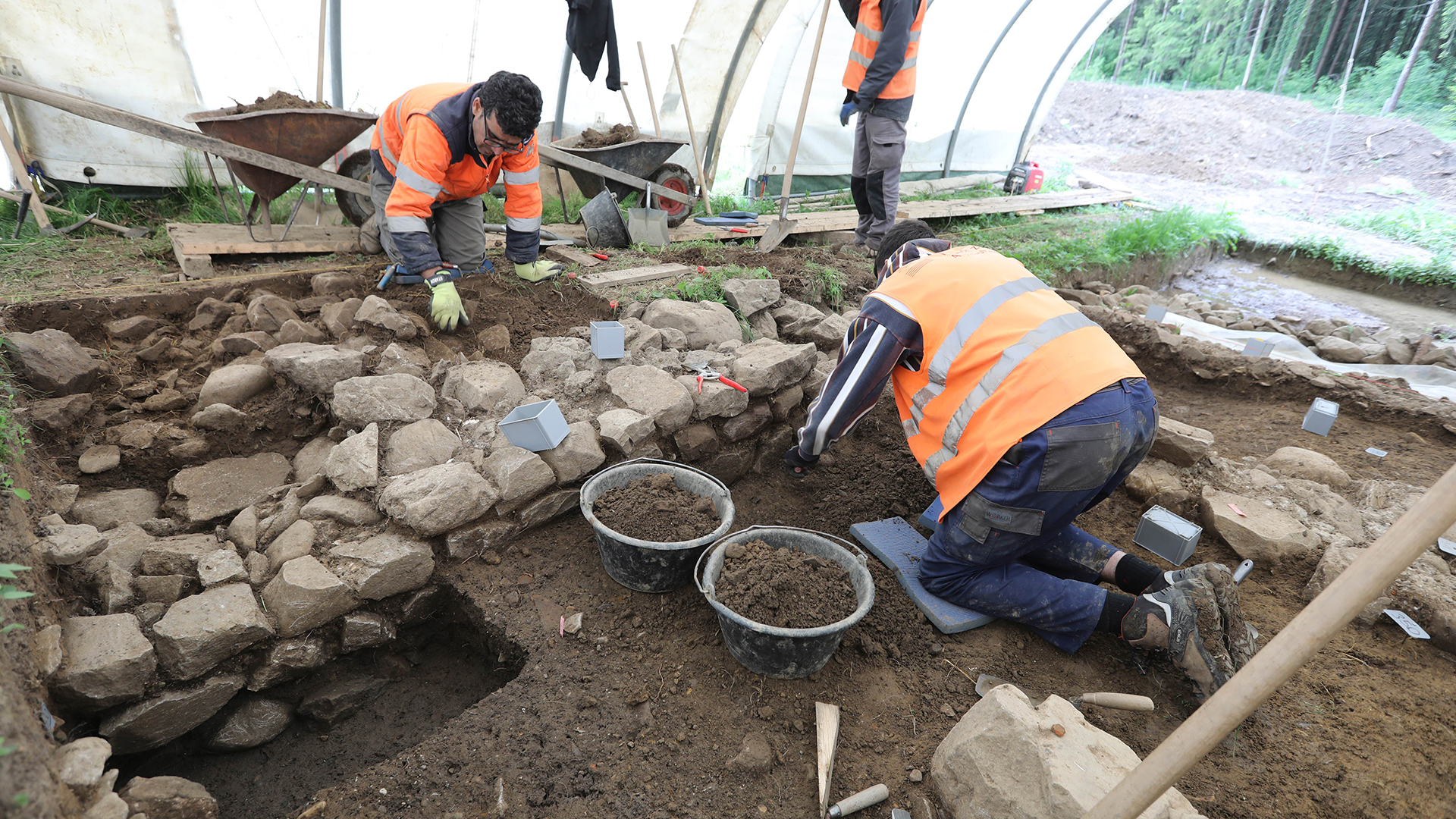 Para arkeolog berjaket visibilitas tinggi sedang menggali tembok rendah.