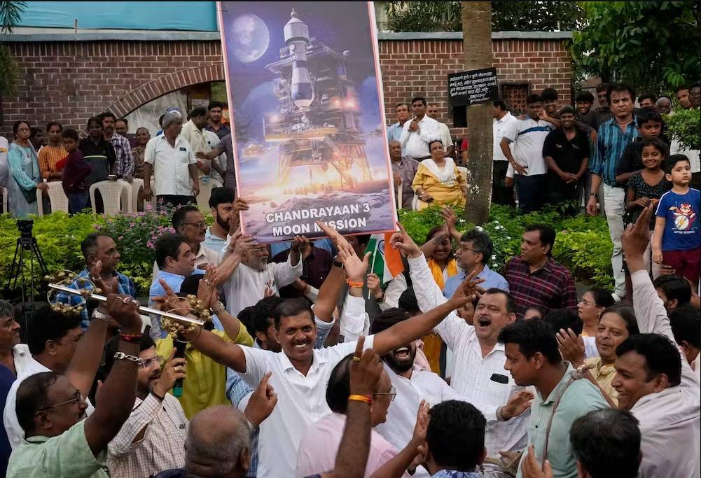 Sekelompok orang yang bersorak dan tersenyum memegang tanda yang menggambarkan pendarat Chandrayaan-3.