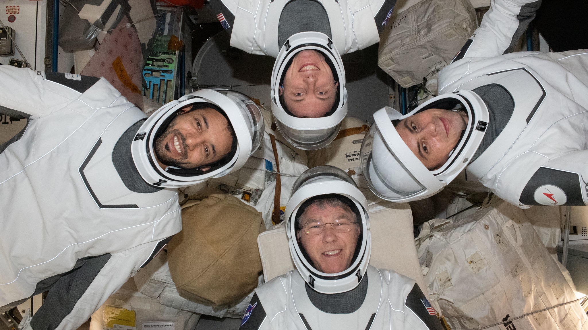 empat astronot mengambang di modul stasiun luar angkasa dengan pakaian antariksa.  mereka terlihat menghadap ke atas dan masing-masing ditempatkan 90 derajat satu sama lain, membentuk lingkaran